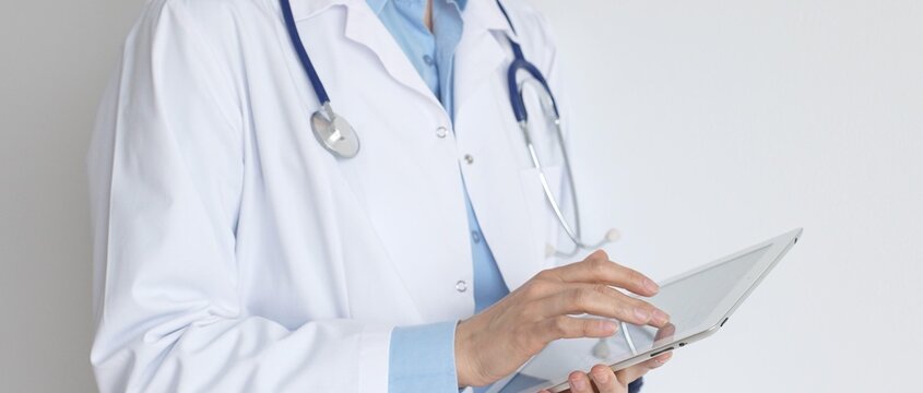 Unknown female doctor, wearing white coat over a blue blouse and a stethoscope, is using a digital tablet, suggesting innovative healthcare practices in medicine. Health care and medicine concept