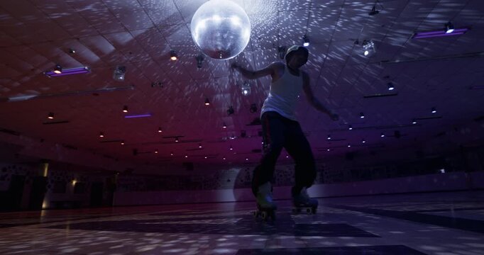 Fun roller skate tricks by man in tank top under sparkling disco ball then falling - low, wide shot