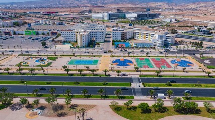 Obraz premium Aerial drone view of a newly renovated modern avenue featuring green public parks, sports courts, palm trees, and urban infrastructure in Agadir, Morocco.
