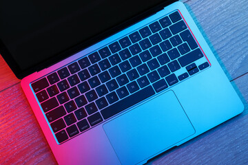 Fototapeta premium Stylish modern laptop in neon lights on wooden table, top view