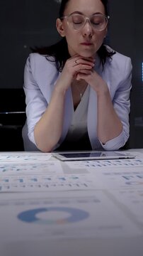 Businesswoman intently reviewing financial charts and reports scattered across her desk while using a digital tablet, working overtime in a corporate environment
