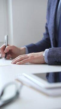 Close up of businessman's hands signing a contract with elegant pen, wearing a blue suit and a wristwatch, sitting at a white desk. Business people concept