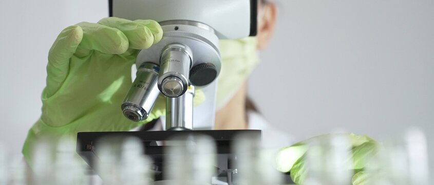 Close up of scientific precision involving green gloved hands adjusting microscope lens, blurred laboratory glassware highlighting medical investigation. Medicine, laboratory and science concept