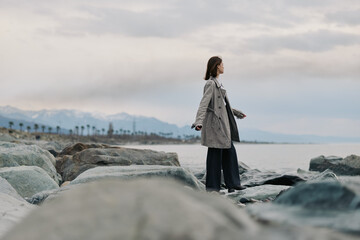A woman in a long coat stands on rocky shore facing the calm sea, mountains in the distance under cloudy sky. Concept of solitude, nature, travel, and peaceful contemplation outdoors. © SHOTPRIME STUDIO