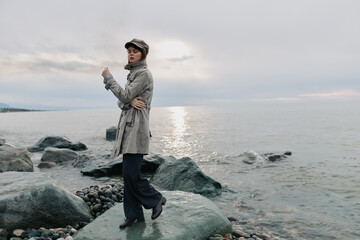 Young woman in coat and hat stands on rocky beach near calm sea during cloudy sunset. Fashion outdoor portrait with serene water background and soft natural light. © SHOTPRIME STUDIO