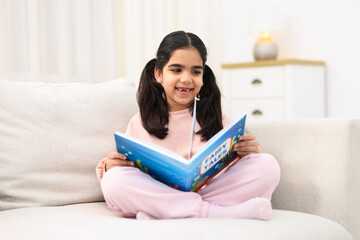 Naklejka premium Cute little girl reading book on couch at home