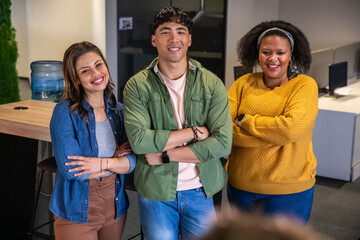 Diverse coworkers posing shoulder-to-shoulder in open office by wooden table and monitors