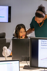Diverse coworkers in officewear working at desk with monitors, lead leaning guiding headset rep