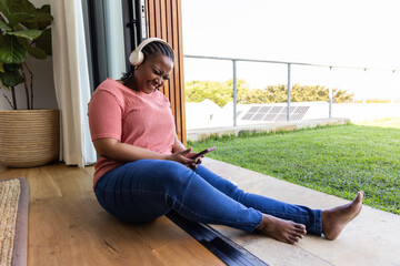 African American woman sitting, laughing while holding phone wearing headset on patio threshold