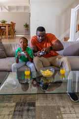 African American dad and son sitting on light-gray sofa holding white controllers, chips and juice © wavebreak3