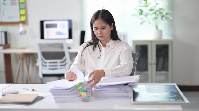 A woman is sitting at a desk with a pile of papers in front of her. She is looking at the papers and seems to be focused on something. Concept of concentration and productivity
