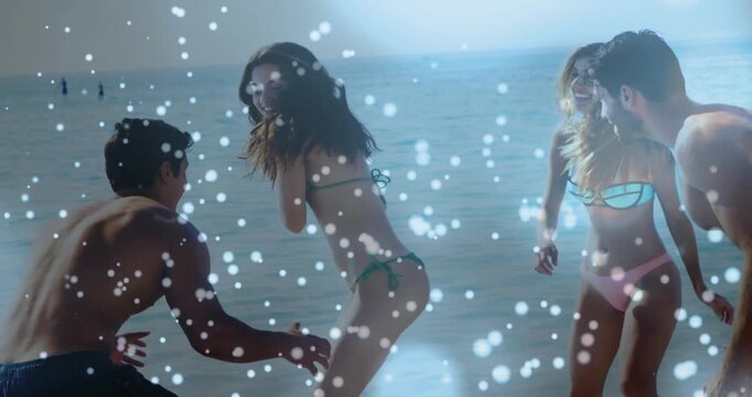 Four friends playing on sandy shore at water's edge wearing swimwear with white ball, woman holding