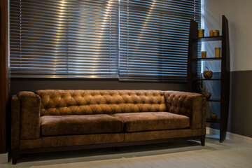 An elegant brown suede Chesterfield sofa sits in a modern office or living room. Light filters through horizontal blinds, creating a calm, sophisticated atmosphere.  © AlfRibeiro