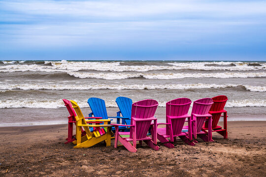 A row of colorful Adirondack chairs arranged in a semicircle on Woodbine Beach in Toronto during a windy spring day with waves.