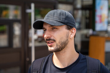 Close-up street portrait of a young man in a black baseball cap and backpack, with beard and hoop earring, looking to the side with a thoughtful expression in soft daylight, blurred urban background © Masarik