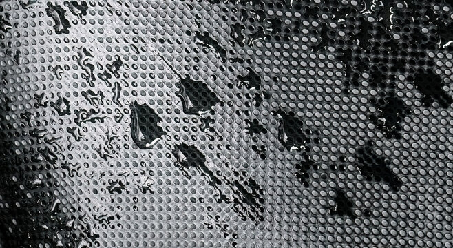 Rainwater on perforated rubber surface with dark abstract pattern. Glossy droplets over industrial mat creating moody textured backdrop. Black tactile flooring with wet reflective details 