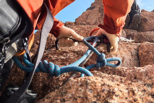 Close-up of mountain climber hands in gloves securing a blue safety rope with knots on a steep rock face during an ascent.