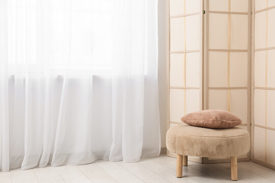 Interior of living room with folding screen, pouf and modern curtains hanging on big window
