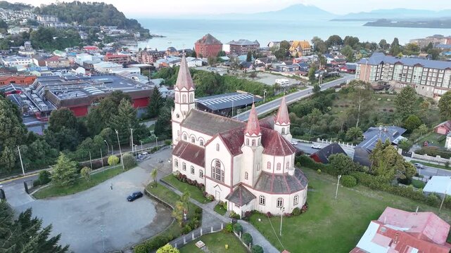 Cinematic aerial circling the iconic Sacred Heart Church (Iglesia del Sagrado Coraz&oacute;n de Jes&uacute;s) in Puerto Varas, Chile, showcasing its red spires and German-influenced architecture against city sky