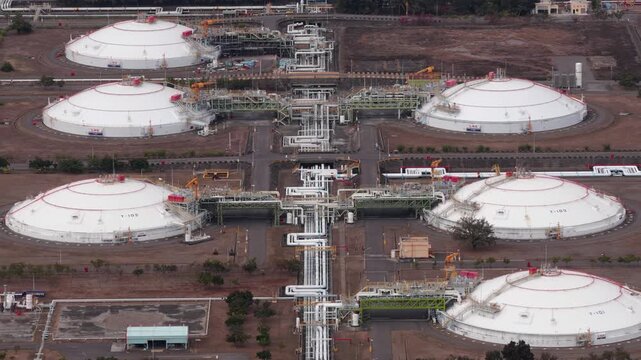 High angle aerial perspective flying over a large liquefied natural gas terminal, showing massive white spherical storage tanks, intricate pipelines, and the surrounding industrial complex