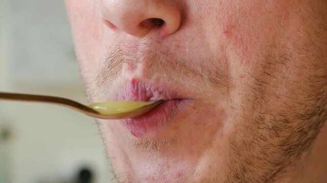 Extreme close up young unshaven man with chapped lips eating sweet, thick, natural honey from golden spoon, savoring delicious and healthy natural sweetener for tasty snack. Man eating sweet honey