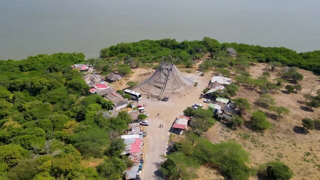 Aerial shot of Totumo Volcano with lush greenery and scattered houses visible. Clear sky above and calm water nearby.