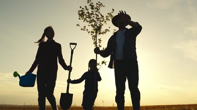 A family of four is planting a tree in a field. The man is holding a shovel and the woman is holding a watering can. The children are also holding shovels and are helping to plant the tree. The scene