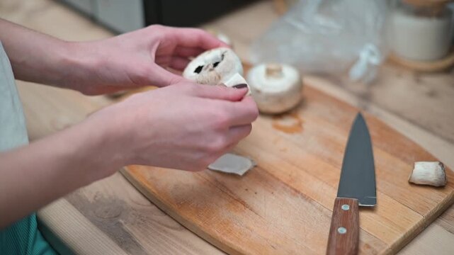 Hands remove the stem from a mushroom and prepare to stuff it with a filling, using a knife on a wooden cutting board
