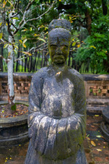 A stone statue of a mandarin stands in the Mausoleum of Emperor Dong Khanh, Hue, Vietnam., a popular tourist destination, The statue is covered in moss and lichen.