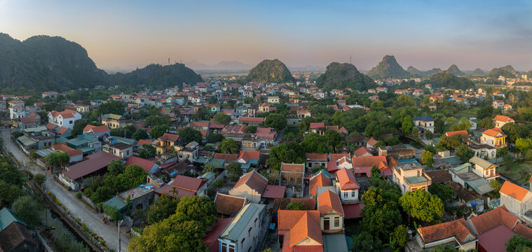A panoramic view of Hoa Lu Ancient Capital, Ninh Binh, Vietnam, showcases the city's unique landscape. Limestone karsts rise above the red-tiled roofs of homes nestled among lush greenery. ,