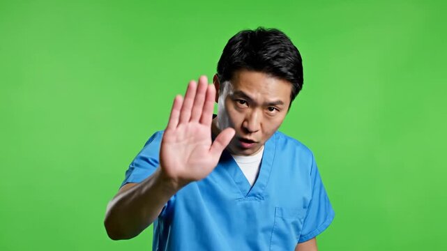Asian Doctor in scrubs on green screen shows stop gesture, health and safety message, and warning