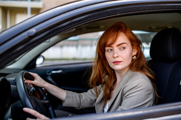 Naklejka premium Serious young redhead woman focused on driving a car through city traffic. Concept of safe driving, road concentration, automotive insurance, and professional female driver commuting in a vehicle.