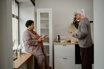 Senior Black woman sitting on windowsill talking to senior Caucasian man cooking in modern kitchen,...