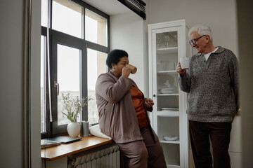 Fototapeta premium Senior Black woman and senior Caucasian man standing in kitchen drinking from mugs, smiling and engaging in friendly conversation near window with natural daylight coming in