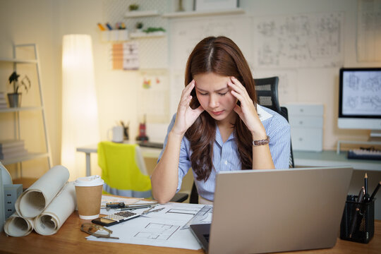 Female architect feeling stressed while working late on building design plans at her office desk.