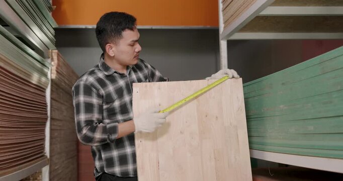 Asian male carpenter holds tape measure across wooden board surface inside woodworking material store, reviewing size specification and preparing materials for interior furniture production process