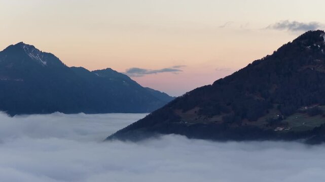 Aerial view of Swiss mountains piercing a thick sea of fog over Lake Walensee. A serene winter sunset glows over the clouds near Walenstadt.