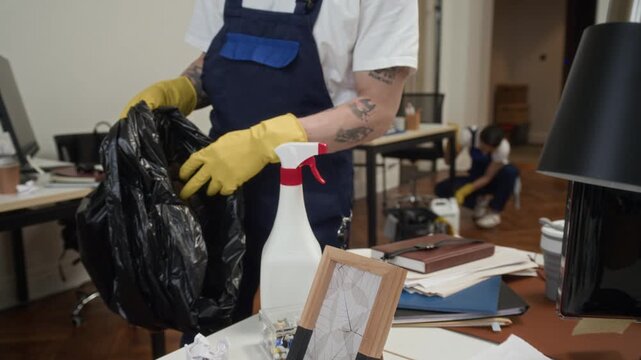Cropped shot of male tattooed janitor in gloves picking up office litter from table and placing in bag while doing cleanup with colleagues
