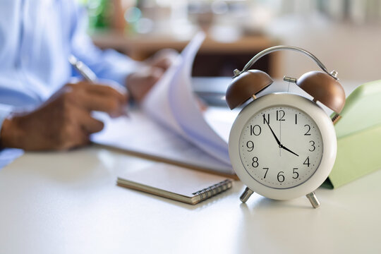 close up at alarm clock 4PM. blurred background asian businessman working in evening with documents checking report and business contract in office before end of work time at 5 PM