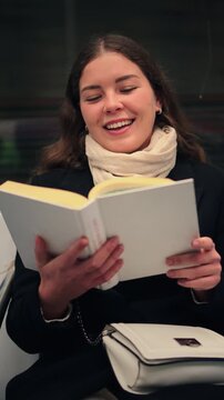 A cheerful young woman is thoroughly enjoying her time as she reads a captivating book in a warm and inviting indoor setting, showcasing her love for literature and relaxation.