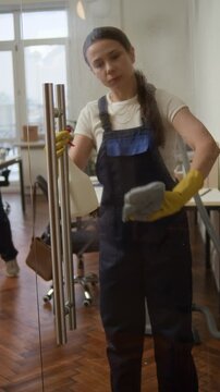 Vertical shot of young Caucasian female cleaner using sprayer and wiping glass door with rag while doing office cleanup with colleagues