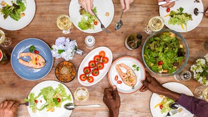 Fototapeta premium Top view of diverse hands enjoy lunch meal with salmon and salad, healthy food. Lunch menu, healthy salad and salmon on wood table, top view photo, diverse people hands enjoy lunch meal, sharing food