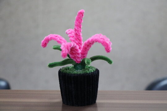 A creative and colorful handmade flower made from pink and green pipe cleaners (chenille stems), displayed in a small black pot on a wooden surface. This artificial plant represents DIY crafts, childh