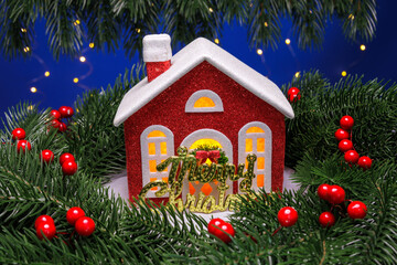 A red Christmas house with letters surrounded by decorated Christmas tree branches on a blue...