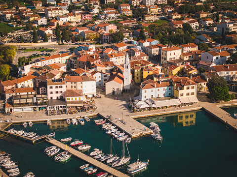 Aerial view of a harbor filled with boats resting near a charming town square dominated by a white church tower, Fazana, Istria County, Croatia.