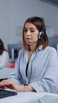 Focused female call center agent wearing headset and working at computer in modern customer support office environment with colleagues in background
