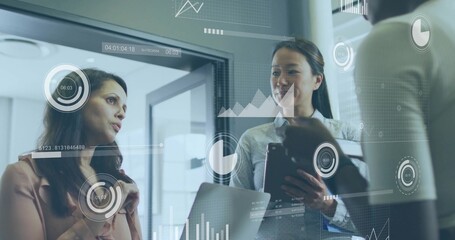 Reviewing three businesswomen examining HUD charts in glass office, in blazers with tablet, laptop