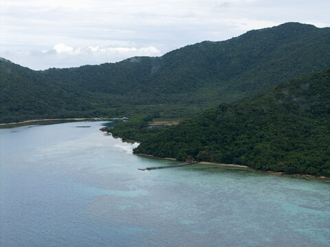 Aerial view of lush green mountains meeting the turquoise sea, a pier extending into the calm waters at Turtle Conservation, Karimunjawa, Central Java, Indonesia.