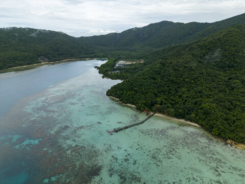 Aerial view of a wooden pier stretching into the turquoise waters meeting lush green hills under a cloudy sky at Turtle Conservation, Karimunjawa, Central Java, Indonesia.