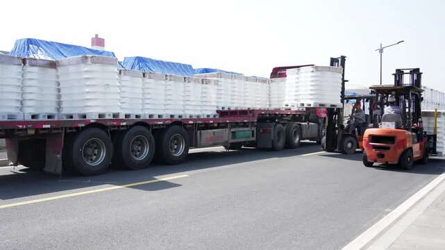 Truck transporting stacked white chemical drums covered with blue tarpaulin at logistics yard